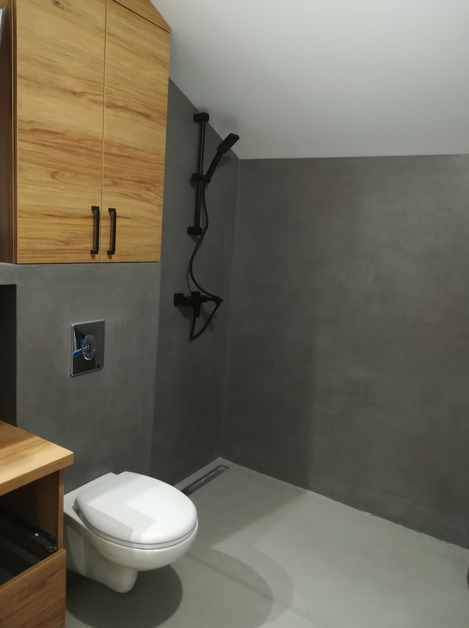 Minimalistyczna łazienka z szarą ścianą, drewnianą szafką i czarną armaturą prysznicową. Uwagę przyciąga nowoczesna toaleta podwieszana.