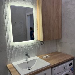 Paweł Jóźwiak - Nowoczesna łazienka z podświetlanym lustrem, umywalką nablatową i pralką pod blatem, wykończona płytkami w dwóch odcieniach szarości.