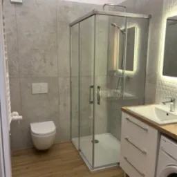 Nowoczesna łazienka z kabiną prysznicową, podwieszaną toaletą, umywalką z szafką i pralką, wykończona szarymi płytkami imitującymi beton i drewnopodobną podłogą.