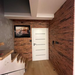 Paweł Jóźwiak - Korytarz z drewnianą podłogą, ceglaną ścianą z obrazem jelenia, białe drzwi z poziomymi frezami i schody z drewnianymi stopniami.