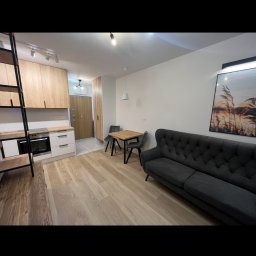 Paweł Jóźwiak - Jasne, przestronne wnętrze mieszkania z drewnianą podłogą, szarą sofą, stołem z krzesłami i aneksem kuchennym z antresolą.