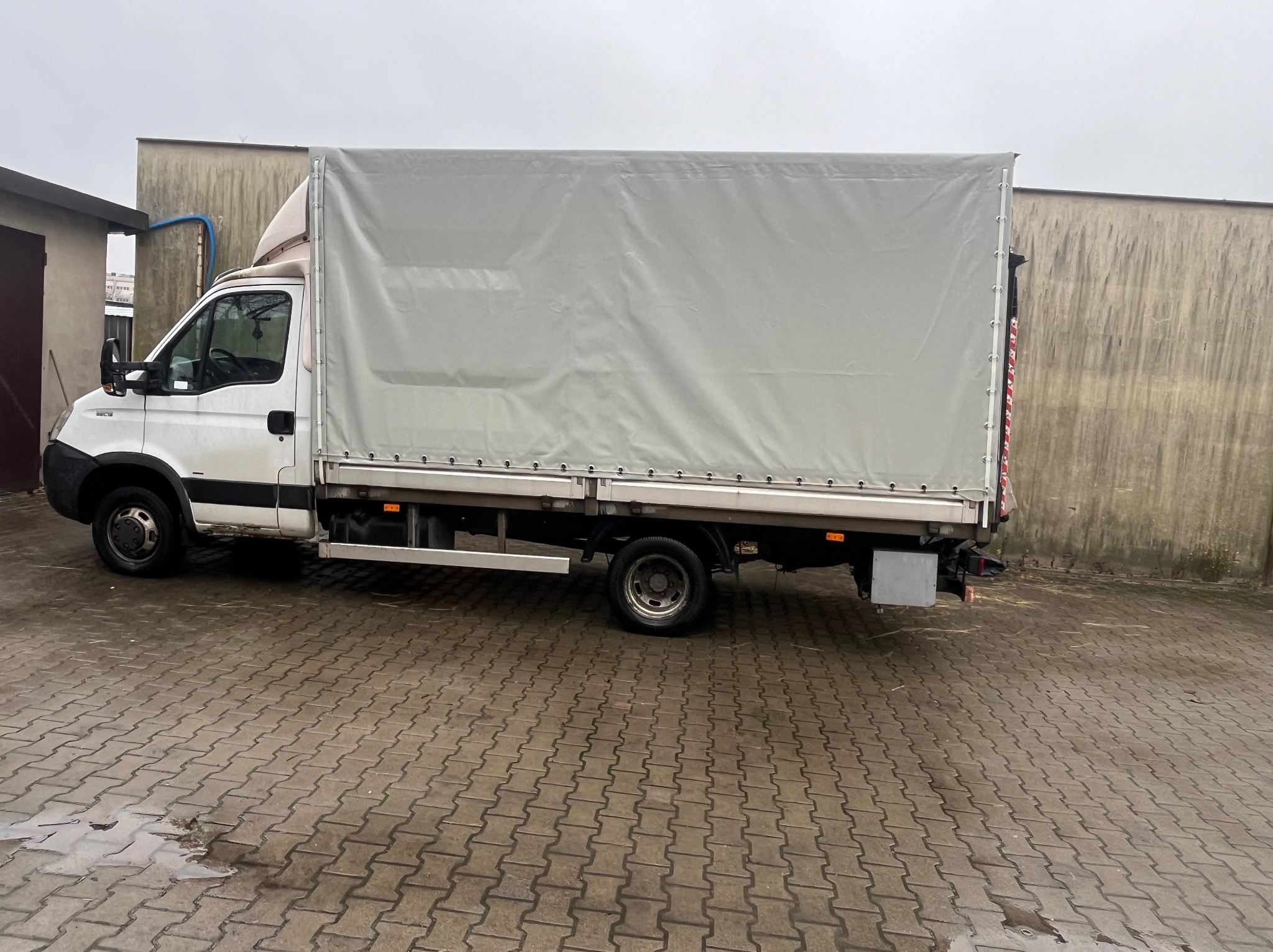 Biały samochód dostawczy z plandeką na mokrym brukowanym podwórku, widoczne ślady użytkowania i zabrudzenia.