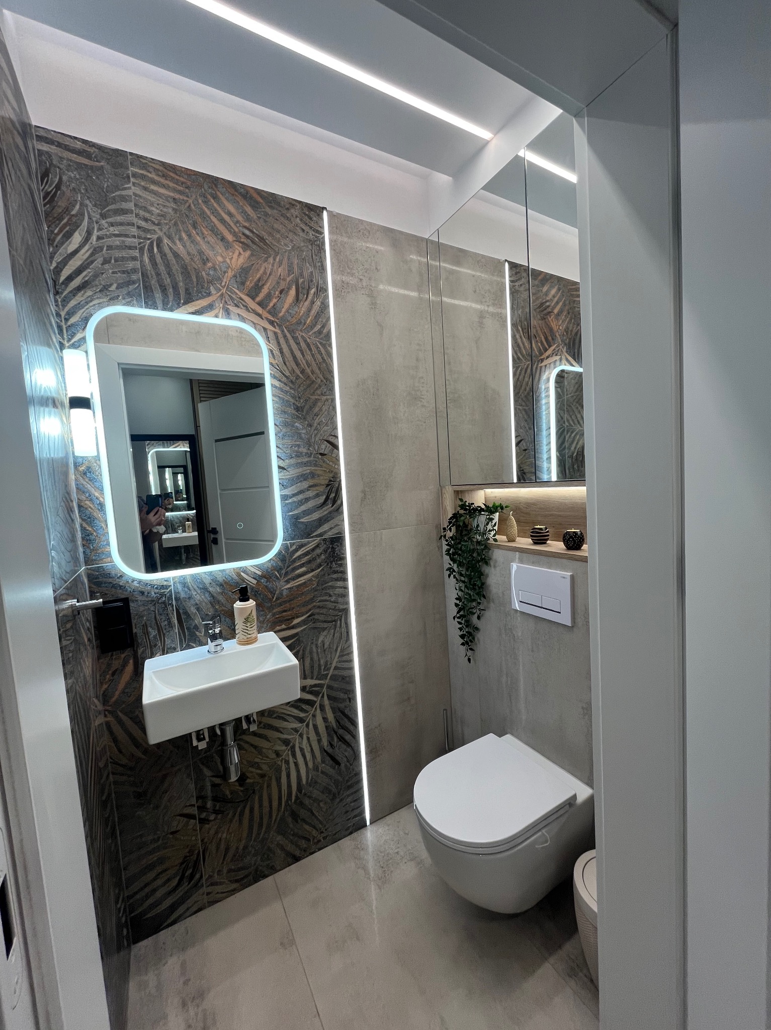 Nowoczesna łazienka z podświetlanym lustrem, umywalką nablatową, wiszącą toaletą i roślinami dekoracyjnymi.