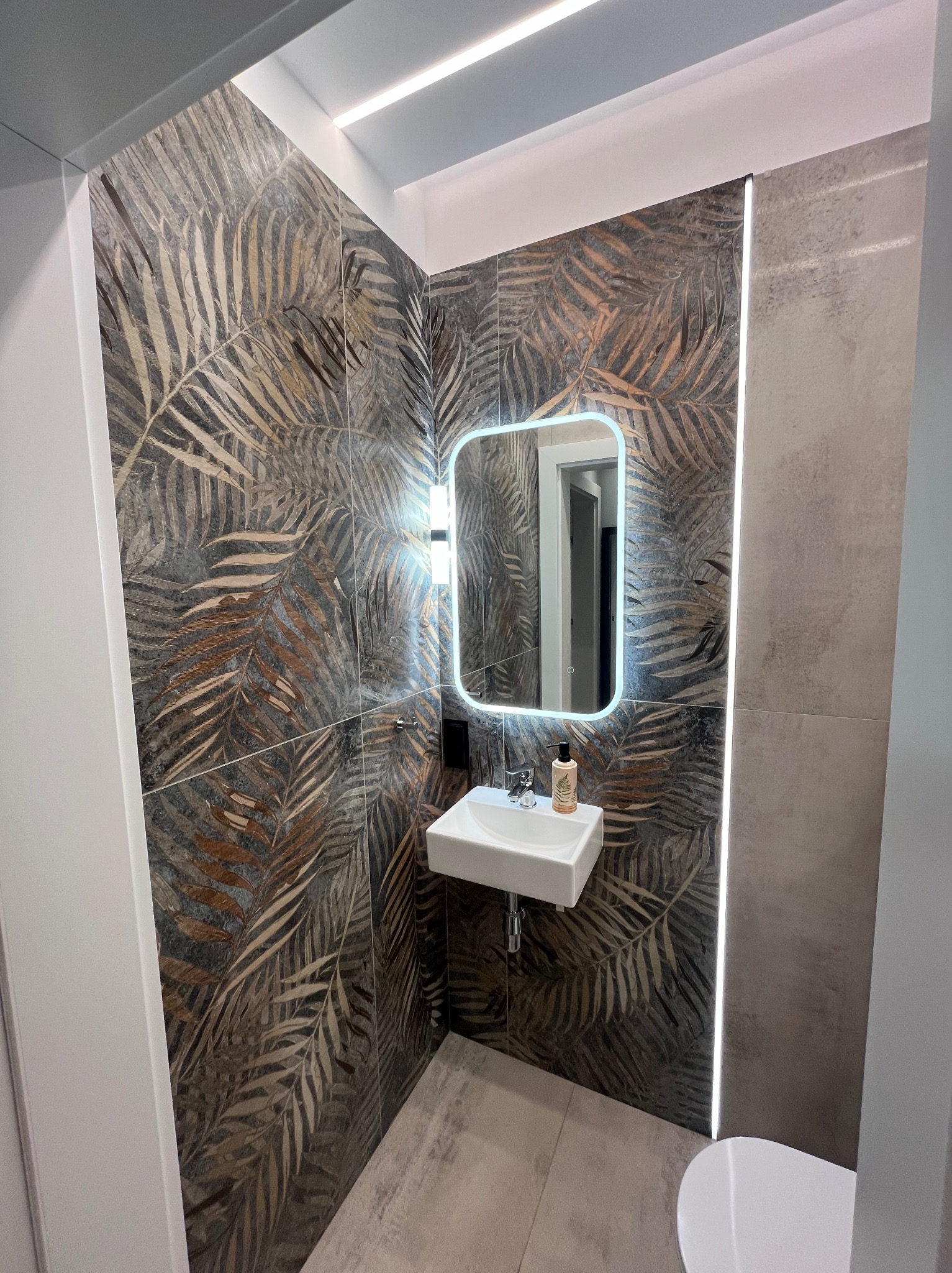 Nowoczesna łazienka z umywalką, podświetlanym lustrem i płytkami z motywem liści palmowych na ścianie.