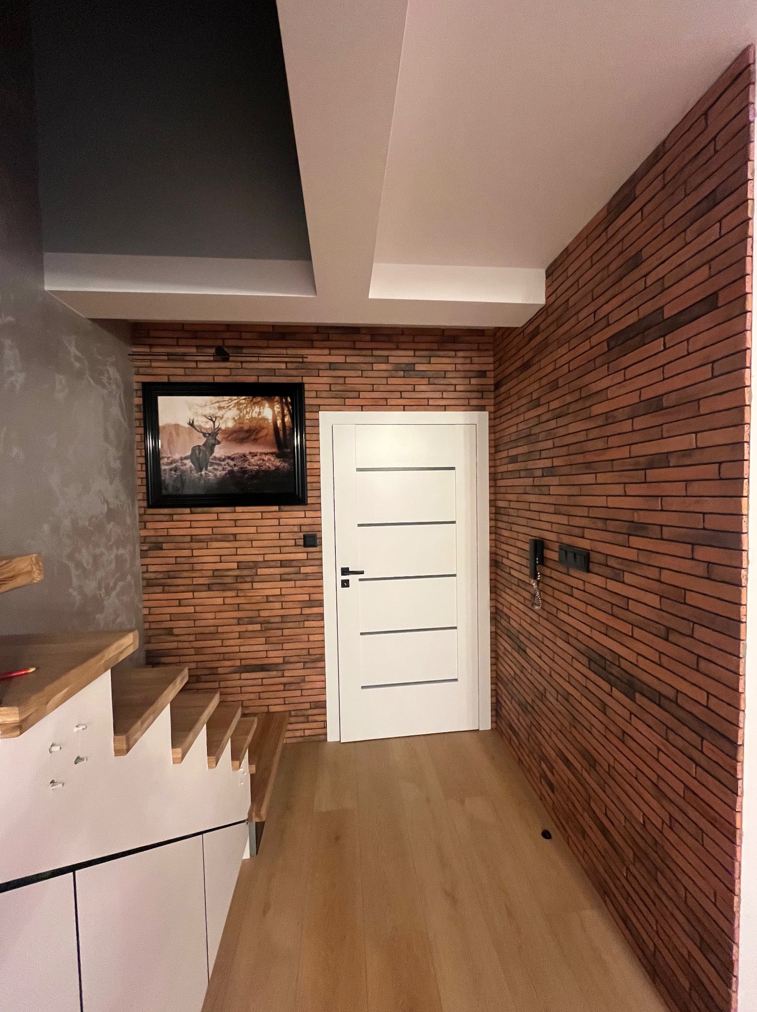 Korytarz z drewnianą podłogą, ceglaną ścianą z obrazem jelenia, białe drzwi z poziomymi frezami i schody z drewnianymi stopniami.