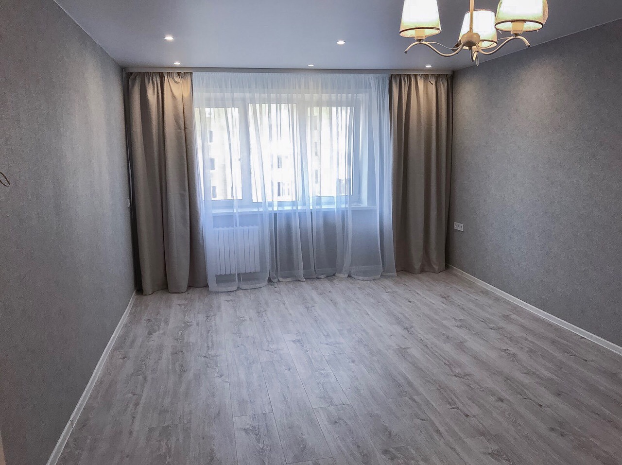 Pusty pokój po remoncie: szare ściany z fakturą, jasna podłoga imitująca drewno, okno z białym grzejnikiem i beżowymi zasłonami, biały sufit z reflektorami i żyrandolem.