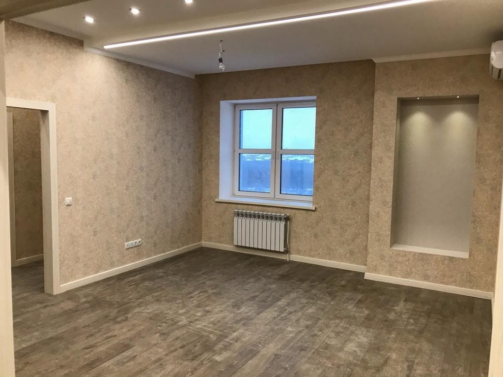 Wykończone wnętrze pokoju z beżowymi tapetami o delikatnym wzorze, panele podłogowe imitujące drewno, białe okno z grzejnikiem oraz wnęka z oświetleniem.