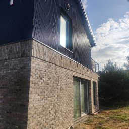 GRUPA POMORSKA PERłA SP. Z O.O. - Nowoczesny dom z elewacją z ciemnej blachy trapezowej i cegły klinkierowej w odcieniach beżu, widok z dołu na narożnik budynku, z widocznymi oknami i fragmentem balkonu.