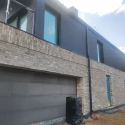 Nowoczesny dom w Gdyni z elewacją z jasnej cegły i czarnym panelem, widok na garaż i okna, teren wokół w trakcie prac wykończeniowych.