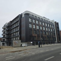 GRUPA POMORSKA PERłA SP. Z O.O. - Nowoczesny, wielopiętrowy budynek z balkonami i ciemną elewacją, widoczny z poziomu ulicy, z elementami placu budowy i graffiti na murze w tle.