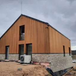 Nowoczesny dom z drewnianą elewacją i ceglaną podmurówką w trakcie wykończenia terenu, widoczna jednostka zewnętrzna klimatyzacji.