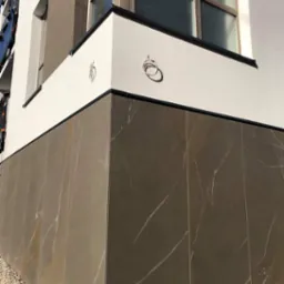 Fragment elewacji budynku z szarymi płytami imitującymi marmur i białym tynkiem, widoczne wyprowadzone kable elektryczne, mały fragment okna i kamyczki przy podstawie ściany.