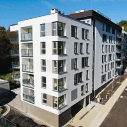 Nowoczesny budynek wielorodzinny z białymi i szarymi elewacjami, balkonami z balustradami ze szkła i nierdzewnej stali, widok z góry na teren w trakcie zagospodarowania.