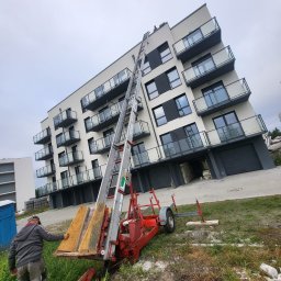 GRUPA POMORSKA PERłA SP. Z O.O. - Podnośnik meblowy ustawiony pod nowoczesnym apartamentowcem z balkonami, przygotowany do transportu materiałów na wyższe piętra, pracownik zabezpiecza ładunek.