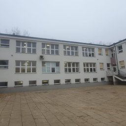 GRUPA POMORSKA PERłA SP. Z O.O. - Dziedziniec szkolny otoczony budynkiem z białymi ścianami i licznymi oknami, widoczne schody zewnętrzne i kostka brukowa na podwórzu, pochmurne niebo.