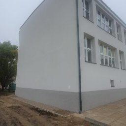 GRUPA POMORSKA PERłA SP. Z O.O. - Odnowiona elewacja budynku z widocznym cokołem w kolorze szarym, świeżo pomalowana na biało, z nowymi oknami i rynną. Teren wokół budynku częściowo uporządkowany, z widocznymi śladami prac...