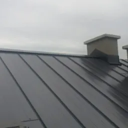 Nowe, grafitowe pokrycie dachowe z blachy na dachu domu, widoczne dwa kominy z szarym tynkiem, pochmurne niebo w tle.