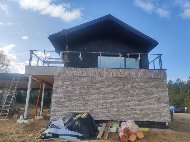 Dom w trakcie budowy z elewacją wykończoną cegłą klinkierową, widoczne rusztowanie, balustrada balkonu w trakcie montażu oraz materiały budowlane na ziemi.