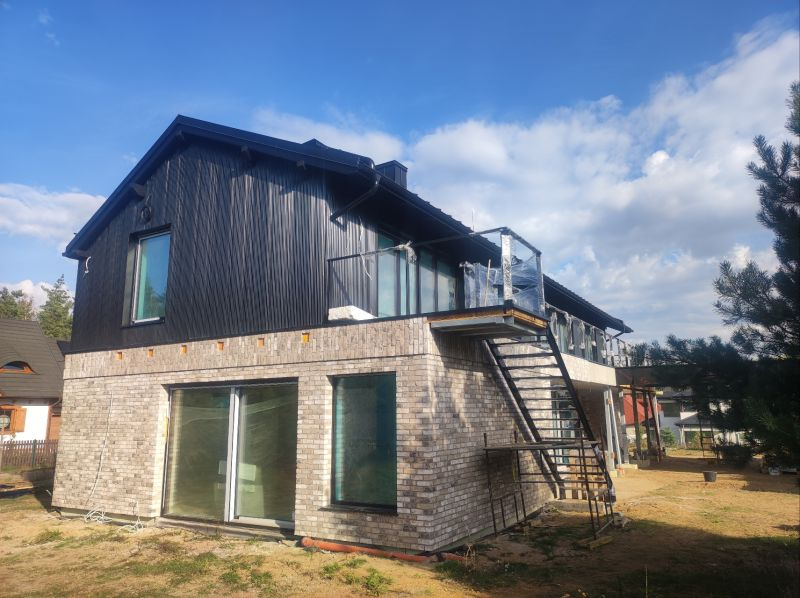 Nowoczesny dom z elewacją z czarnego drewna i cegły, z zewnętrznymi schodami prowadzącymi na balkon, częściowo zabezpieczonym folią ochronną.