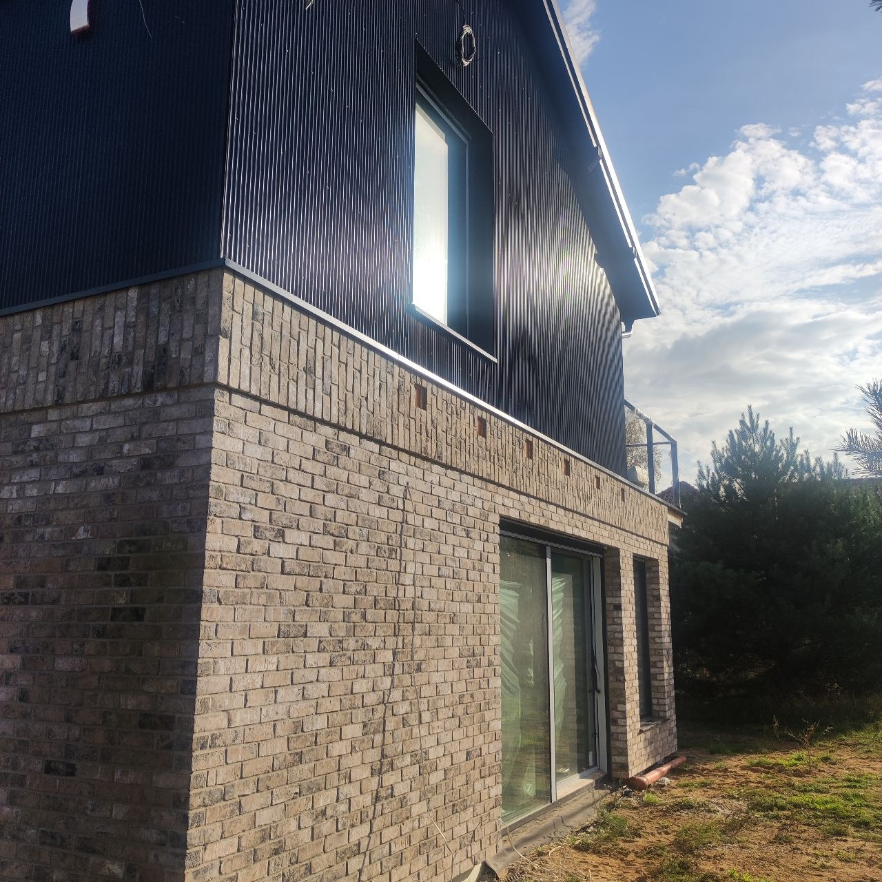 Nowoczesny dom z elewacją z ciemnej blachy trapezowej i cegły klinkierowej w odcieniach beżu, widok z dołu na narożnik budynku, z widocznymi oknami i fragmentem balkonu.