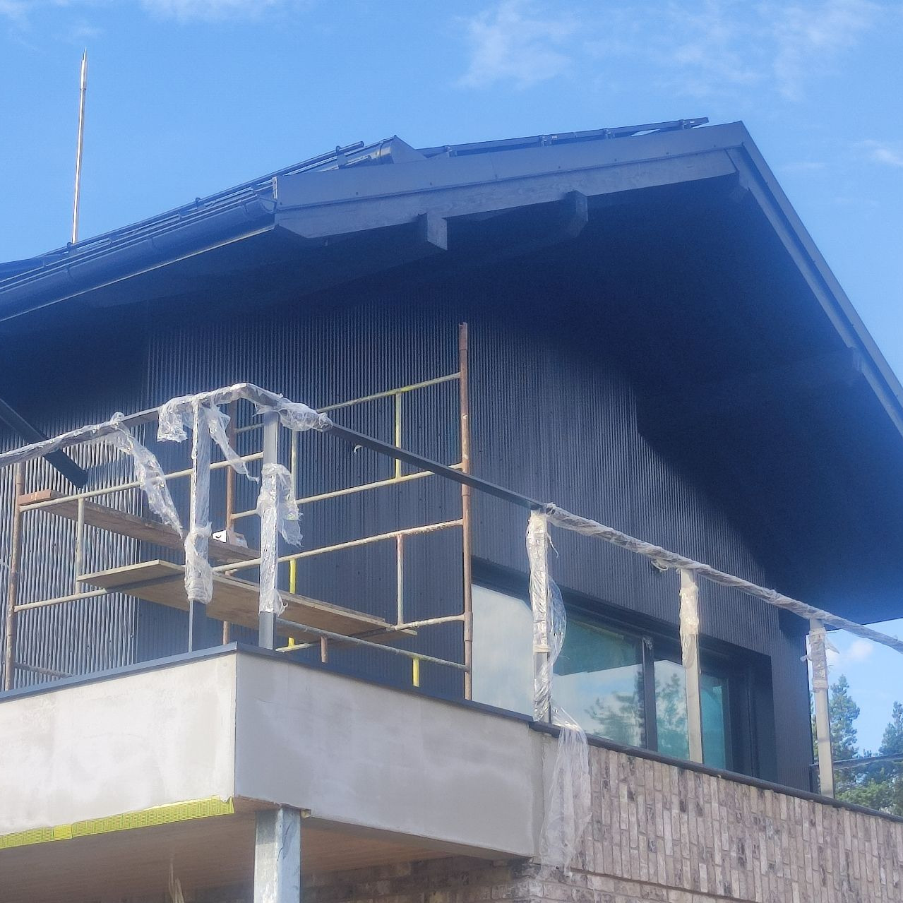 Elewacja domu z blachy trapezowej w kolorze grafitowym, częściowo zakryta rusztowaniem i folią ochronną, widoczny fragment balkonu z surowym tynkiem.