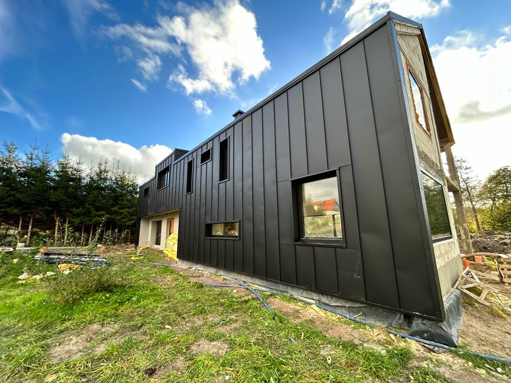 Nowoczesny dom w trakcie wykończenia z czarną elewacją z blachy na rąbek stojący, widoczne fragmenty surowego muru i okna, zielony trawnik i błękitne niebo z chmurami w tle.