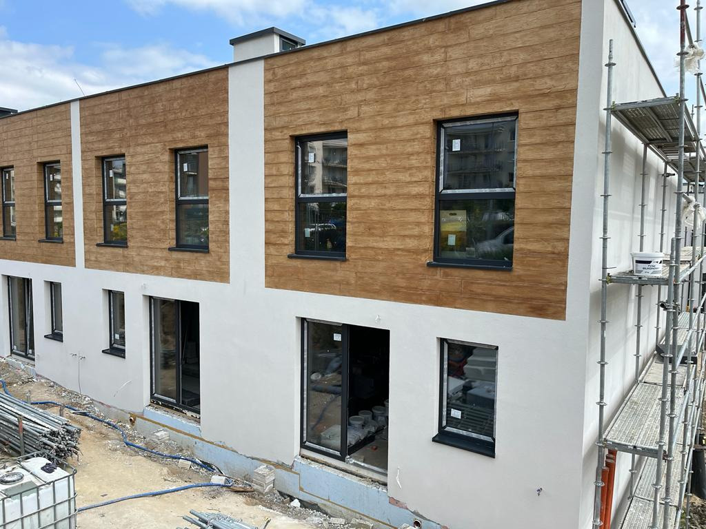 Dwukondygnacyjny budynek z elewacją w kolorze białym i brązowym imitującym drewno, rusztowanie po prawej stronie, widoczne rury i pojemniki na ziemi sugerujące trwające prace wykończeniowe.