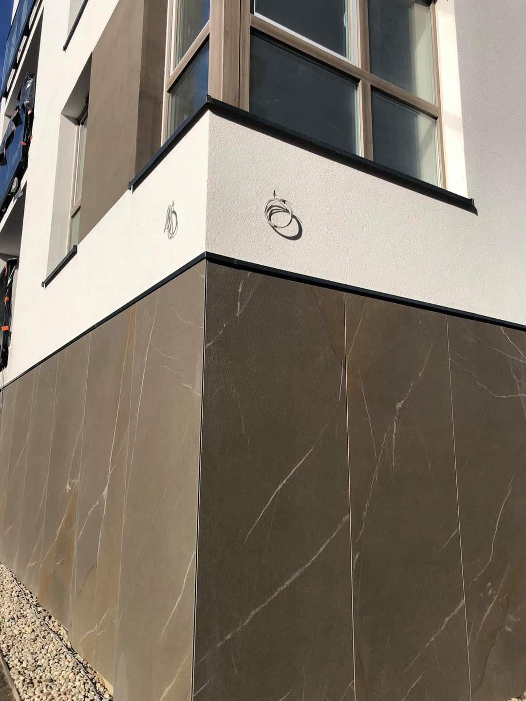 Fragment elewacji budynku z szarymi płytami imitującymi marmur i białym tynkiem, widoczne wyprowadzone kable elektryczne, mały fragment okna i kamyczki przy podstawie ściany.