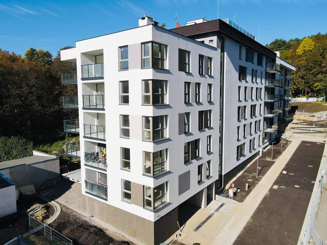 Nowoczesny budynek wielorodzinny z białymi i szarymi elewacjami, balkonami z balustradami ze szkła i nierdzewnej stali, widok z góry na teren w trakcie zagospodarowania.