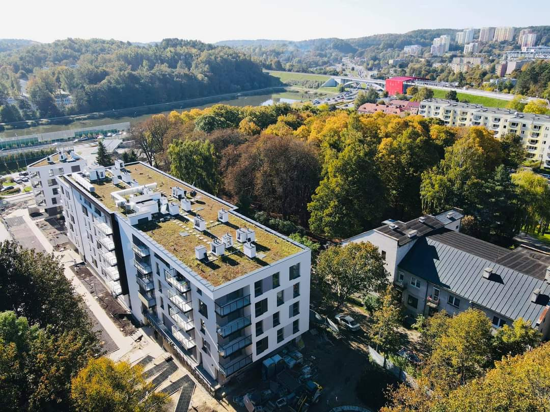 Nowoczesny budynek mieszkalny z zielonym dachem widziany z góry, z balkonami i systemem wentylacyjnym na dachu, w tle park, rzeka i zabudowa miejska.