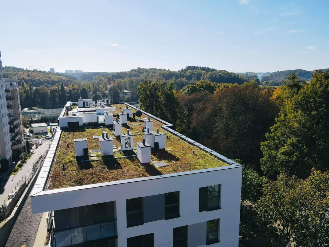 Widok z góry na nowoczesny budynek z zielonym dachem porośniętym roślinnością, z widocznymi elementami wentylacji i kominami, na tle lasu i zabudowy miejskiej w oddali.