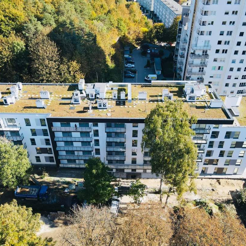Nowoczesny budynek wielorodzinny z zielonym dachem widziany z góry, z widoczną osobą na balkonie i elementami zagospodarowania terenu wokół.