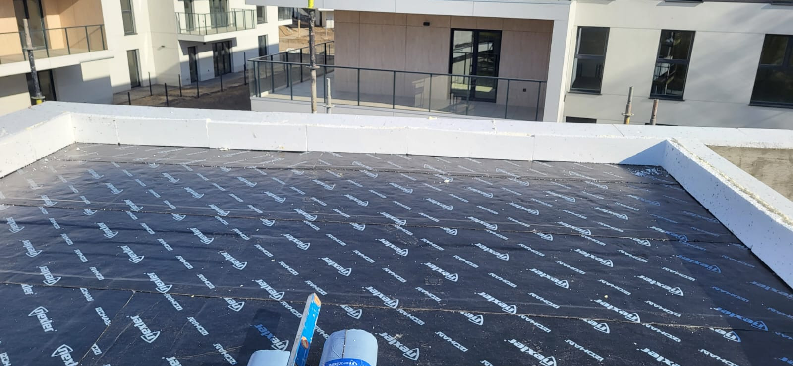 Izolacja termiczna płaskiego dachu styropianem z hydroizolacją z papy termozgrzewalnej, widoczne narzędzia i materiały budowlane, budynki wielorodzinne w tle.