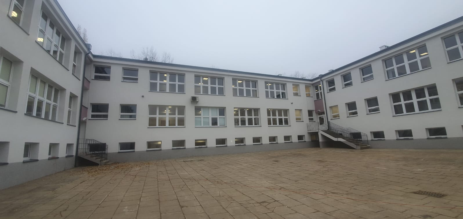 Dziedziniec szkolny otoczony budynkiem z białymi ścianami i licznymi oknami, widoczne schody zewnętrzne i kostka brukowa na podwórzu, pochmurne niebo.