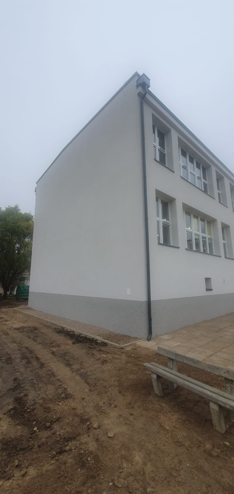 Odnowiona elewacja budynku z widocznym cokołem w kolorze szarym, świeżo pomalowana na biało, z nowymi oknami i rynną. Teren wokół budynku częściowo uporządkowany, z widocznymi śladami prac...