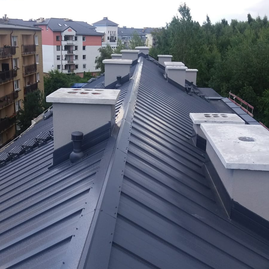 Nowoczesny, ciemnoszary dach z blachy trapezowej z kilkoma betonowymi kominami na tle miejskiego krajobrazu.