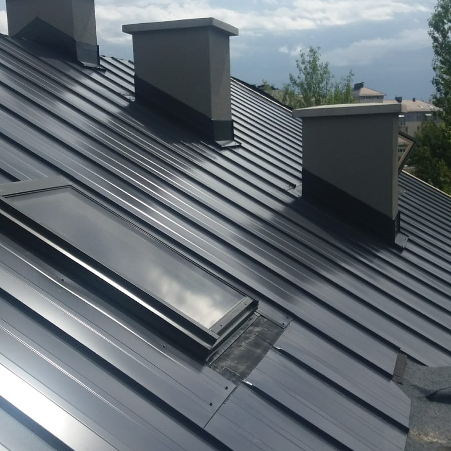 Szary metalowy dach z oknem dachowym i trzema kominami na tle pochmurnego nieba i zabudowy miejskiej.