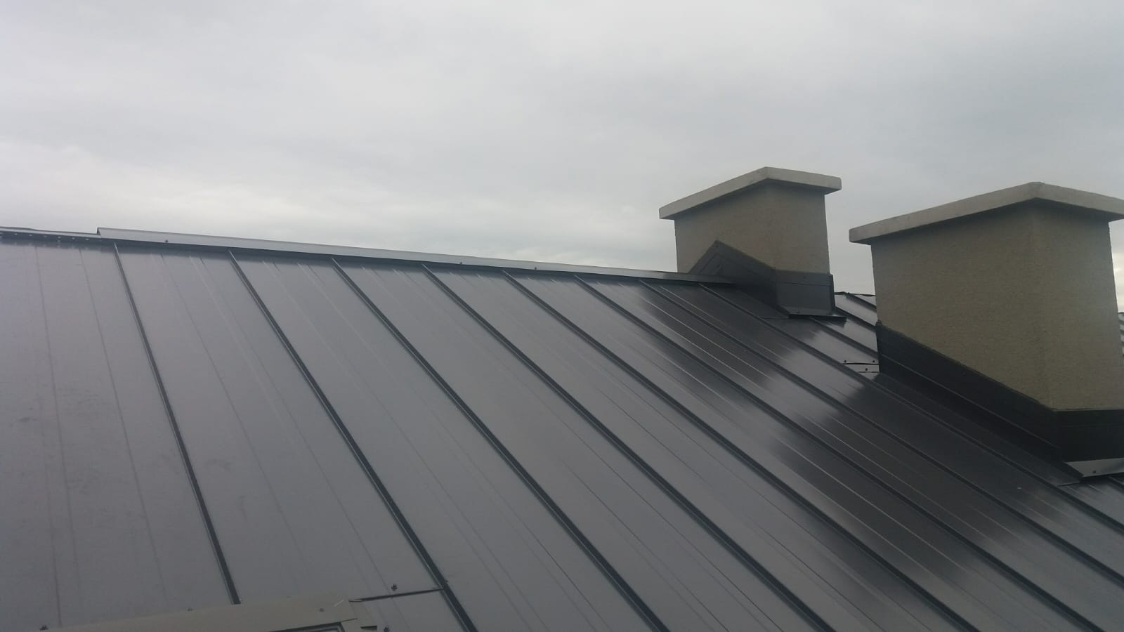 Nowe, grafitowe pokrycie dachowe z blachy na dachu domu, widoczne dwa kominy z szarym tynkiem, pochmurne niebo w tle.