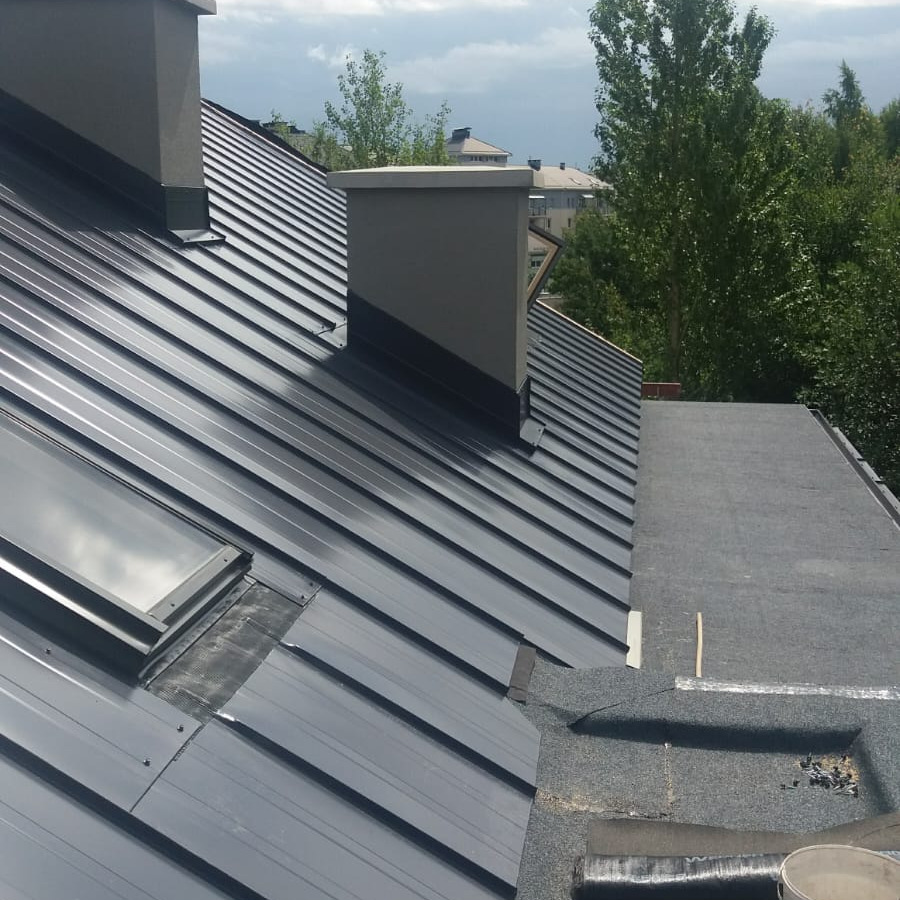 Szary dach z blachy trapezowej z dwoma kominami i oknem dachowym, widok z góry, w tle zieleń i budynki.