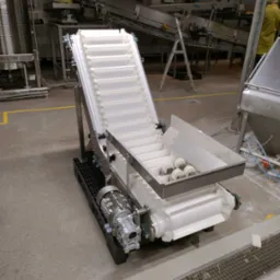 Biały, modułowy przenośnik taśmowy z silnikiem elektrycznym, ustawiony pod kątem, w hali produkcyjnej z widocznymi innymi urządzeniami i żółtymi liniami na podłodze.