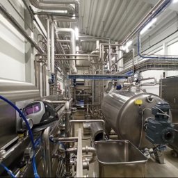 SPAW WARS - Kompleksowa instalacja przemysłowa z rurami ze stali nierdzewnej i zbiornikami, widoczna maska spawalnicza, oświetlenie sufitowe w hali produkcyjnej.