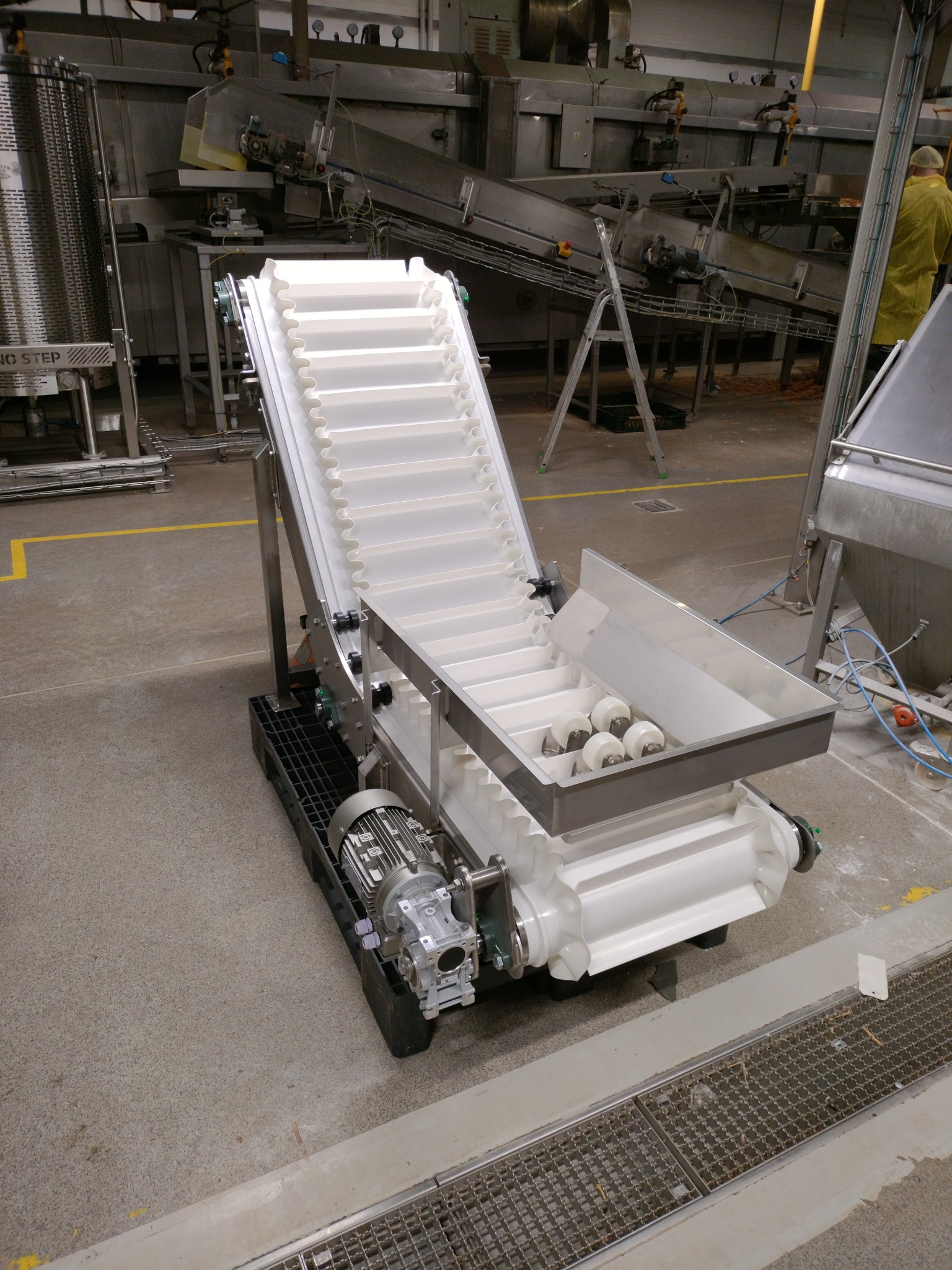 Biały, modułowy przenośnik taśmowy z silnikiem elektrycznym, ustawiony pod kątem, w hali produkcyjnej z widocznymi innymi urządzeniami i żółtymi liniami na podłodze.