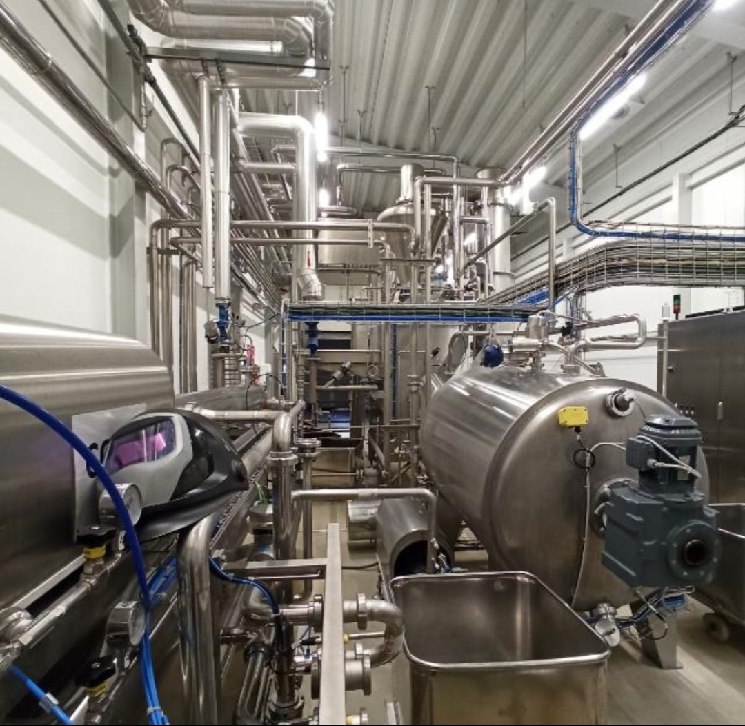 Kompleksowa instalacja przemysłowa z rurami ze stali nierdzewnej i zbiornikami, widoczna maska spawalnicza, oświetlenie sufitowe w hali produkcyjnej.