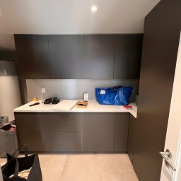 Artur Hinc - Nowoczesna łazienka z szarymi szafkami, białym blatem, beżowymi płytkami podłogowymi i niebieską torbą na blacie; widoczne drobne przedmioty i akcesoria.