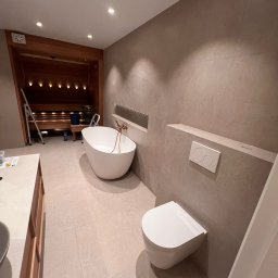 Artur Hinc - Nowoczesna łazienka z wolnostojącą wanną, podwieszaną toaletą i sauną w tle, wykończona płytkami w odcieniach beżu, z elementami miedzianej armatury.