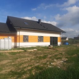 Artur Hinc - Nowo wybudowany dom jednorodzinny z czarnym dachem, elewacją w kolorze białym i pomarańczowym, okna zabezpieczone folią, widoczne zbiorniki na wodę przy ścianie, trawnik z przodu, budynki...