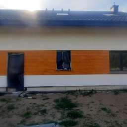 Artur Hinc - Elewacja domu jednorodzinnego w trakcie malowania, z widocznym pasem w kolorze pomarańczowym na środku, okna i drzwi zabezpieczone folią, widoczny fragment dachu z dachówką.