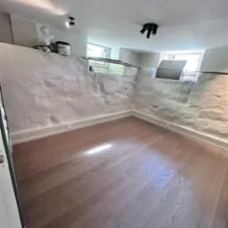 Wnętrze piwnicy po renowacji z nową podłogą z paneli, surowe ściany pomalowane na biało, widoczne okna piwniczne i oświetlenie punktowe.