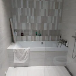 Nowoczesna łazienka z wanną obłożoną beżowymi płytkami imitującymi marmur, szklany parawan nawannowy, deszczownica i półka w ścianie z kosmetykami.