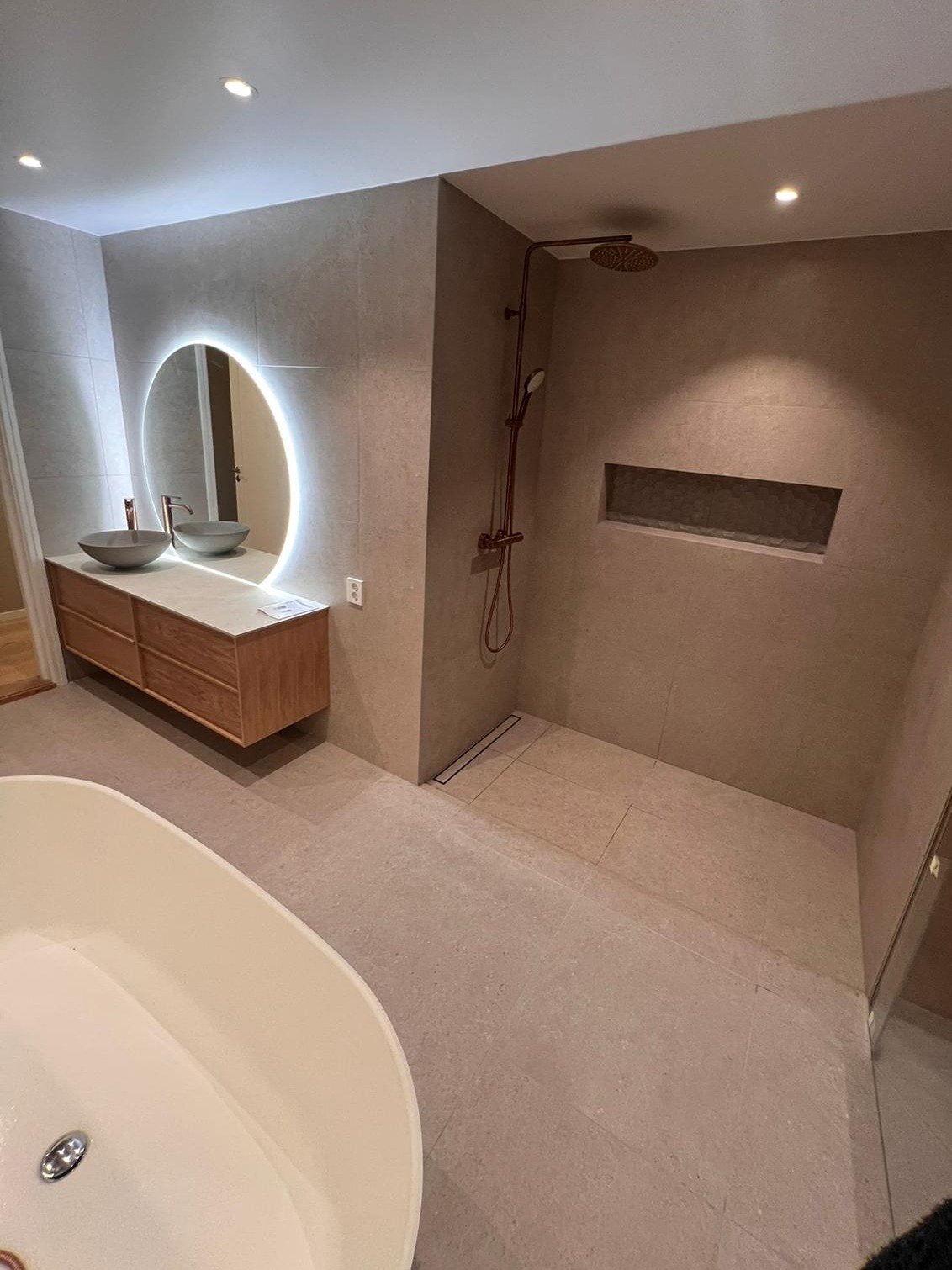 Nowoczesna łazienka z beżowymi płytkami, owalną wanną, drewnianą szafką pod umywalką z okrągłym podświetlanym lustrem oraz prysznicem z deszczownicą w kolorze miedzi.